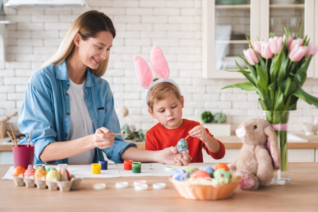 Happy positive mother and little son decorating eggs for Easter at home